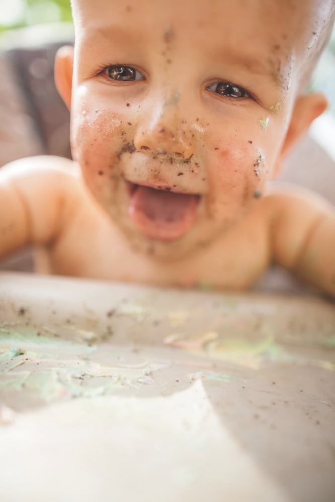 A Naked One Year-Old Baby Boy Covered in Birthday Cake Sits in a High Chair and Sticks His Tongue Out